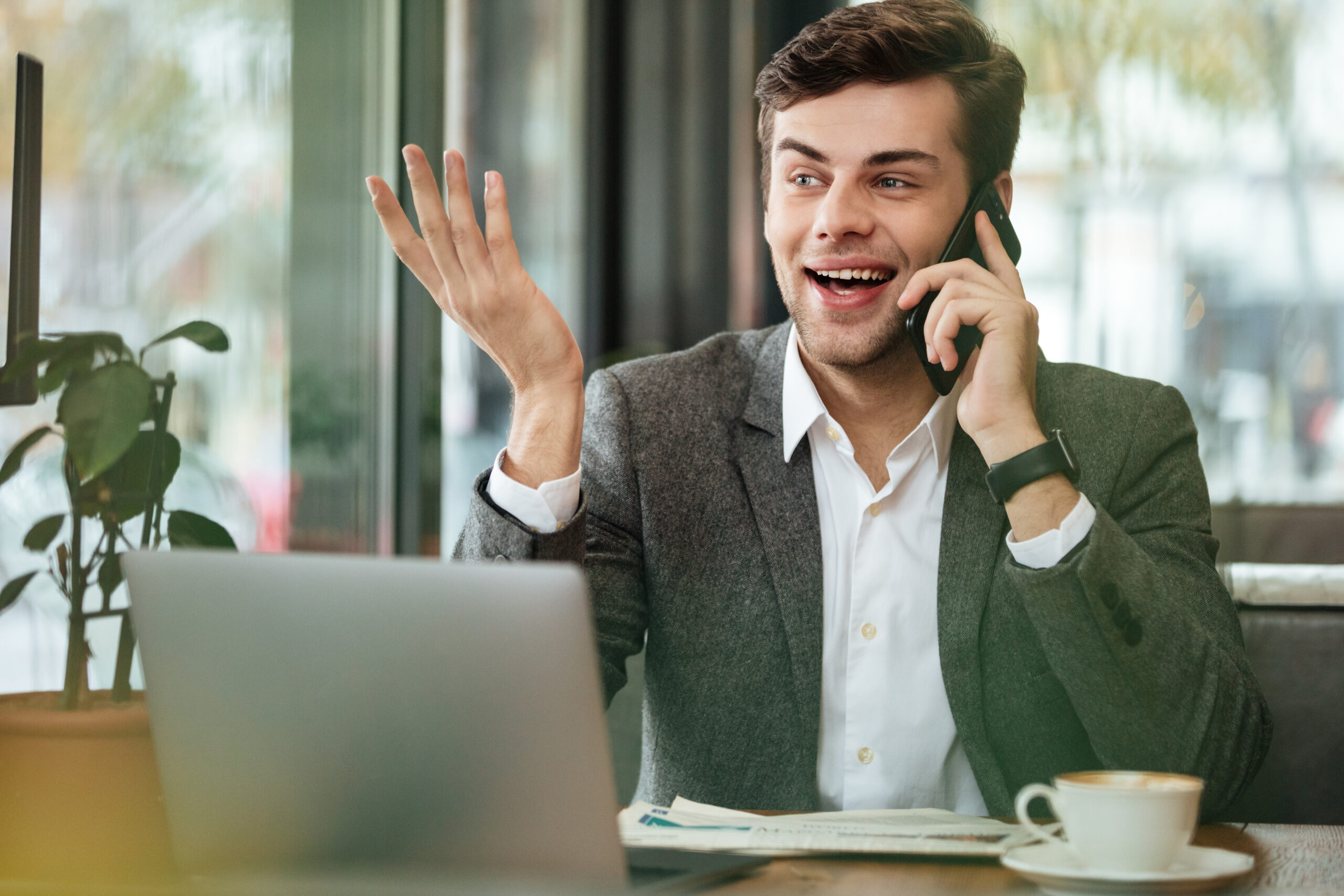 Happy business man sitting by the table in cafe with laptop computer and talking by smartphone while looking away